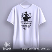 Футболка тато білого кольору, зображена з фронтального ракурсу на вішалці, фон світло-сірий, принт чорного кольору з написом 'БУТИ ТАТОМ ОЗНАЧАЄ БУТИ ГЕРОЄМ ДЛЯ СВОЇХ ДІТЕЙ КОЖНОГО ДНЯ' та зображенням батька з дитиною на плечах.