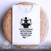 Футболка тато білого кольору, складена на плетеній основі, фон світлий, принт чорного кольору з написом 'БУТИ ТАТОМ ОЗНАЧАЄ БУТИ ГЕРОЄМ ДЛЯ СВОЇХ ДІТЕЙ КОЖНОГО ДНЯ' та зображенням батька з дитиною на плечах.