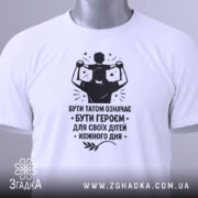 Футболка тато білого кольору, складена на поверхні, фон світло-сірий, принт чорного кольору з написом 'БУТИ ТАТОМ ОЗНАЧАЄ БУТИ ГЕРОЄМ ДЛЯ СВОЇХ ДІТЕЙ КОЖНОГО ДНЯ' та зображенням батька з дитиною на плечах, з зеленим ярликом.