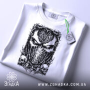 Футболка темна маска, біла, складена на поверхні, з ракурсу зверху, фон світло-сірий, принт з чорним малюнком маски та черепами, на футболці є етикетка з зеленим шнурком.