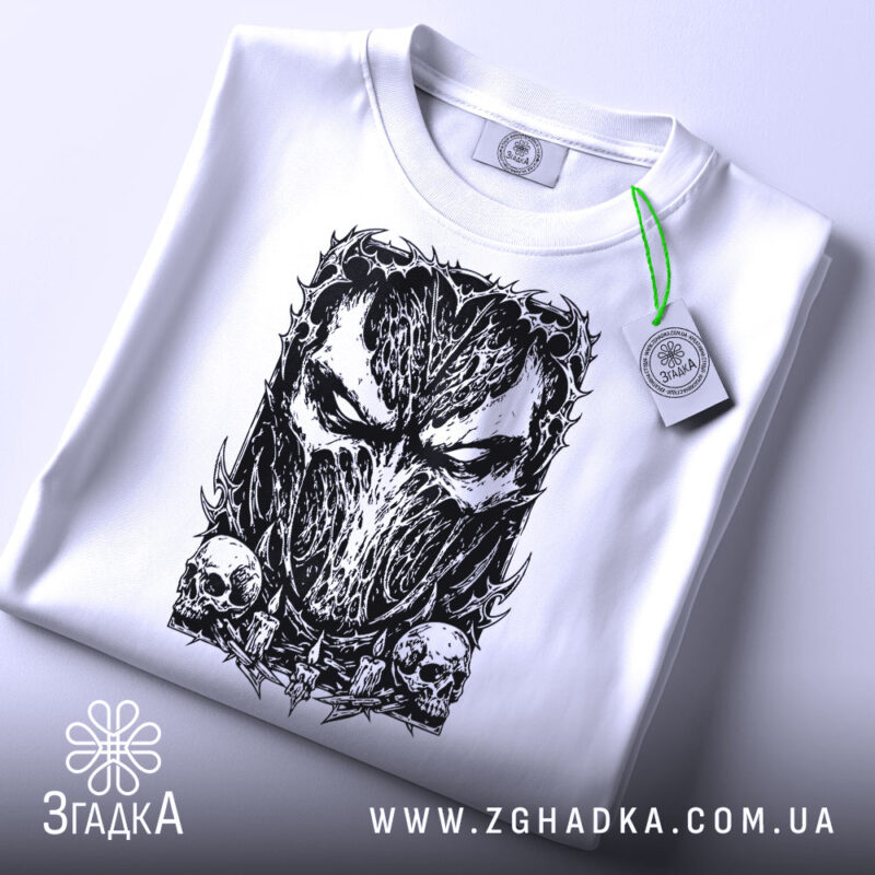 Футболка темна маска, біла, складена на поверхні, з ракурсу зверху, фон світло-сірий, принт з чорним малюнком маски та черепами, на футболці є етикетка з зеленим шнурком.