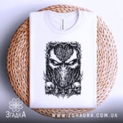 Футболка темна маска, біла, складена на плетеній кошику, фон світло-сірий, принт з чорним малюнком маски та черепами, видно етикетку на горловині.