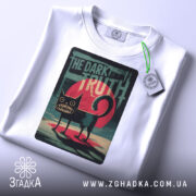 Футболка темна правда, біла, складена на столі, фон світло-сірий, принт з чорним котом на червоному фоні з написом 'THE DARK TRUTH', на комірці бирка з зеленим шнурком.