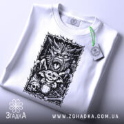 Футболка темний демон, біла, зображена на столі, ракурс зверху, фон світло-сірий, принт з чорними деталями, на якому зображений демон, ведмедик і черепи, з етикеткою зеленого кольору.