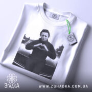 Футболка темний романтик, біла, зображена в плоскому вигляді, з принтом чорного персонажа, що робить жест серця, на фоні автомобіля, з етикеткою зеленого кольору.