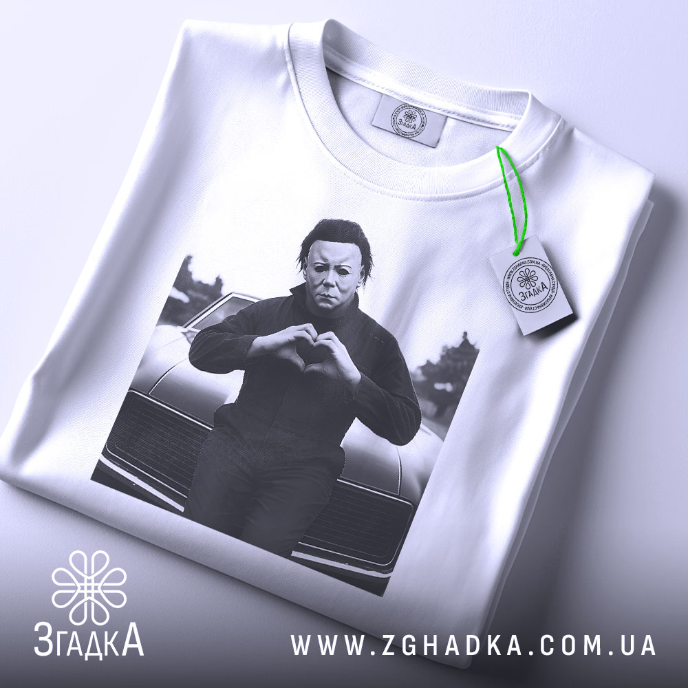 Футболка темний романтик, біла, зображена в плоскому вигляді, з принтом чорного персонажа, що робить жест серця, на фоні автомобіля, з етикеткою зеленого кольору.