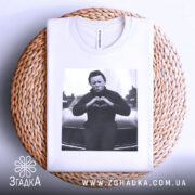 Футболка темний романтик, біла, складена на плетеній основі, з принтом чорного персонажа, що робить жест серця, на фоні автомобіля.