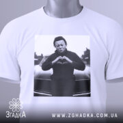 Футболка темний романтик, біла, зображена в плоскому вигляді, з принтом чорного персонажа, що робить жест серця, на фоні автомобіля, з етикеткою зеленого кольору.