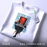 Футболка темний символізм, біла, складена на столі, фон світло-сірий, принт з червоним демоном, що тримає табличку з написом 'WHO ARE YOU?' на чорному фоні, текст на футболці у зелених тонах, з етикеткою з зеленим шнурком.