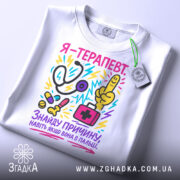 Футболка терапевт білого кольору, складена на плоскій поверхні, фон світло-сірий, принт кольоровий, зображає стетоскоп, пальця вказівника, медичну сумку та інші медичні предмети, текст на футболці українською мовою, прикріплена етикетка з логотипом.