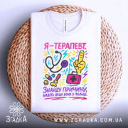 Футболка терапевт білого кольору, складена на плоскій поверхні, фон світло-сірий, принт кольоровий, зображає стетоскоп, пальця вказівника, медичну сумку та інші медичні предмети, текст на футболці українською мовою, розташована на плетеній основі.