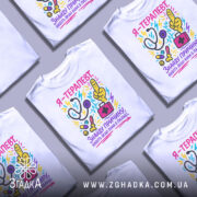 Футболка терапевт білого кольору, зображена на манекені з правого боку, фон світло-сірий, принт кольоровий, містить малюнки стетоскопа, пальця вказівника, медичної сумки та інших медичних предметів, текст на футболці українською мовою.