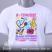 Футболка терапевт білого кольору, складена на плоскій поверхні, фон світло-сірий, принт кольоровий, зображає стетоскоп, пальця вказівника, медичну сумку та інші медичні предмети, текст на футболці українською мовою, прикріплена етикетка з логотипом.