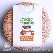 Футболка токсик білого кольору, зображена зверху на плетеному кошику, фон білий, принт з птахом у сонцезахисних окулярах, що тримає каву, текст зеленого та оранжевого кольорів.