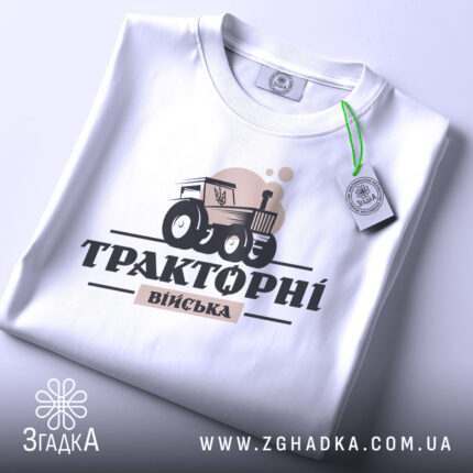 Футболка тракторні війська білого кольору, складена на поверхні, з видимим етикеткою, фон світло-сірий, принт з трактором та написом 'Тракторні війська' в чорному та бежевому кольорах.