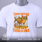 Футболка Trulimero Trulicina білого кольору, складена на столі, фон світло-сірий, принт з котом у сонцезахисних окулярах і риб'ячим тілом, оточений зеленими рослинами, текст 'TRULIMERO TRULICINA' яскраво-оранжевого кольору, на футболці є етикетка з зеленим шнурком.