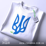 Футболка тризуб голуб миру, біла, зображена на столі, ракурс зверху, фон світло-сірий, принт у вигляді тризуба синього кольору з голубом, що тримає гілочку оливи, з етикеткою на шнурку.