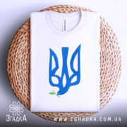 Футболка тризуб голуб миру, біла, зображена на плетеній кошику, ракурс зверху, фон світло-сірий, принт у вигляді тризуба синього кольору з голубом, що тримає гілочку оливи.