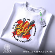 Футболка Тризуб і кайдани, біла, зображена зверху, на складеній футболці, фон світло-сірий, принт з червоним фоном, жовтим тризубом і кайданами, текст чорного кольору, з етикеткою зеленого кольору.