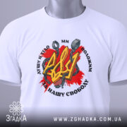 Футболка Тризуб і кайдани, біла, зображена зверху, на складеній футболці, фон світло-сірий, принт з червоним фоном, жовтим тризубом і кайданами, текст чорного кольору, з етикеткою зеленого кольору.