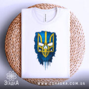 Футболка тризуб білого кольору, зображена в горизонтальному положенні на плетеній кошику, фон світлий, принт тризуба жовтого кольору на синьому фоні, деталі принта яскраві.