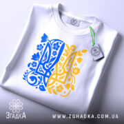 Футболка тризуб український орнамент, біла, зображена зверху, складена на столі, фон світло-сірий, принт у синьо-жовтих тонах з квітковими елементами, на комірці є етикетка.