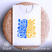 Футболка тризуб український орнамент, біла, зображена зверху на плетеній кошику, фон світло-сірий, принт у синьо-жовтих тонах з квітковими елементами, видна етикетка на комірці.