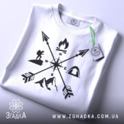 Футболка турист білого кольору, складена на поверхні, зображена з верхнього ракурсу, фон світлий, принт чорного кольору з символами активного відпочинку, на ярлику зелена стрічка.