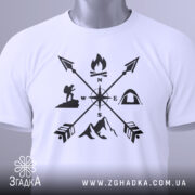 Футболка турист білого кольору, складена на поверхні, зображена з верхнього ракурсу, фон світлий, принт чорного кольору з символами активного відпочинку, на ярлику зелена стрічка.