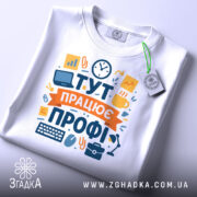 Футболка тут працює профі. Біла футболка, складена на столі, з видимим принтом. Зображення з верхнього ракурсу, фон світло-сірий, на футболці є етикетка з зеленим шнурком.