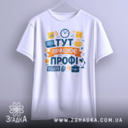 Футболка тут працює профі. Біла футболка з коротким рукавом, підвішена на вішалці, фон світло-сірий. Принт яскравий, з елементами, що символізують роботу, такими як комп'ютер і чашка.