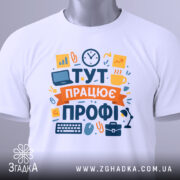 Футболка тут працює профі. Біла футболка, складена на столі, з видимим принтом. Зображення з верхнього ракурсу, фон світло-сірий, на футболці є етикетка з зеленим шнурком.
