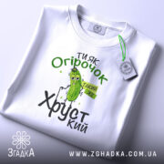 Футболка ти як огірочок, біла, зображена зверху, на фоні світло-сірого кольору, принт з огірком та написом "Ти як огірочок, такий же хрусткий" в зелених та чорних кольорах, з етикеткою на рукаві.