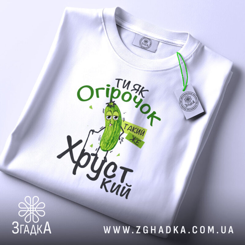 Футболка ти як огірочок, біла, зображена зверху, на фоні світло-сірого кольору, принт з огірком та написом "Ти як огірочок, такий же хрусткий" в зелених та чорних кольорах, з етикеткою на рукаві.