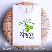 Футболка ти як огірочок, біла, зображена зверху на плетеній кошику, на фоні світлого кольору, принт з огірком та написом "Ти як огірочок, такий же хрусткий" в зелених та чорних кольорах.