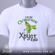 Футболка ти як огірочок, біла, зображена зверху, на фоні світло-сірого кольору, принт з огірком та написом "Ти як огірочок, такий же хрусткий" в зелених та чорних кольорах, з етикеткою на рукаві.