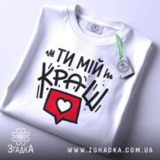 Футболка ти мій краш, білого кольору, зображена в складеному вигляді, фон світло-сірий, принт з текстом "ТИ МІЙ КРАШ" у чорному кольорі та червоний коментар з серцем, на комірці є етикетка.