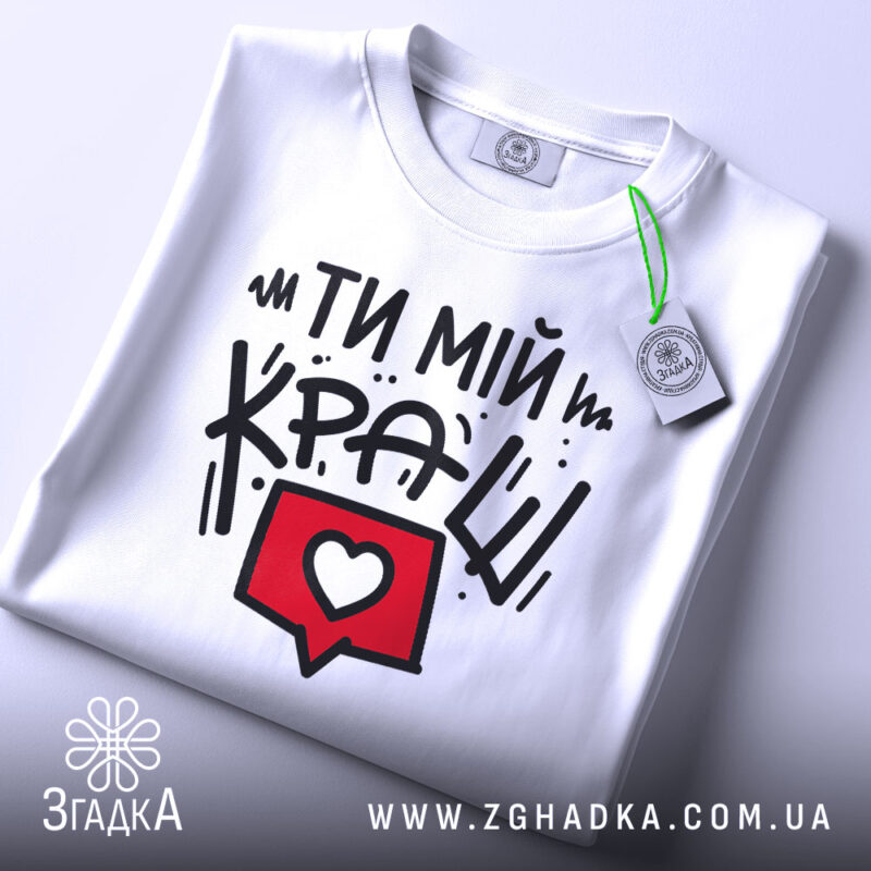 Футболка ти мій краш, білого кольору, зображена в складеному вигляді, фон світло-сірий, принт з текстом "ТИ МІЙ КРАШ" у чорному кольорі та червоний коментар з серцем, на комірці є етикетка.