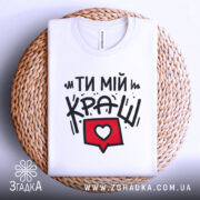 Футболка ти мій краш, білого кольору, зображена в складеному вигляді на плетеній кошику, фон світло-сірий, принт містить текст "ТИ МІЙ КРАШ" у чорному кольорі та червоний коментар з серцем.