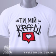 Футболка ти мій краш, білого кольору, зображена в складеному вигляді, фон світло-сірий, принт з текстом "ТИ МІЙ КРАШ" у чорному кольорі та червоний коментар з серцем, на комірці є етикетка.