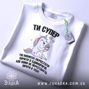Футболка ти супер, біла, зображена зверху, фон світлий, принт з єдинорогом, який тримає торт, текст чорного кольору, на комірці є етикетка.