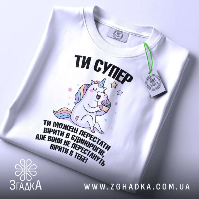 Футболка ти супер, біла, зображена зверху, фон світлий, принт з єдинорогом, який тримає торт, текст чорного кольору, на комірці є етикетка.