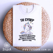 Футболка ти супер, біла, зображена зверху на плетеній основі, фон світлий, принт з єдинорогом, який тримає торт, текст чорного кольору.