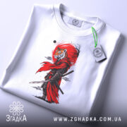 Футболка Тигр самурай воїн, біла, зображена на столі, фон світло-сірий, принт з тигром у червоному плащі, на футболці є етикетка з зеленим шнурком.