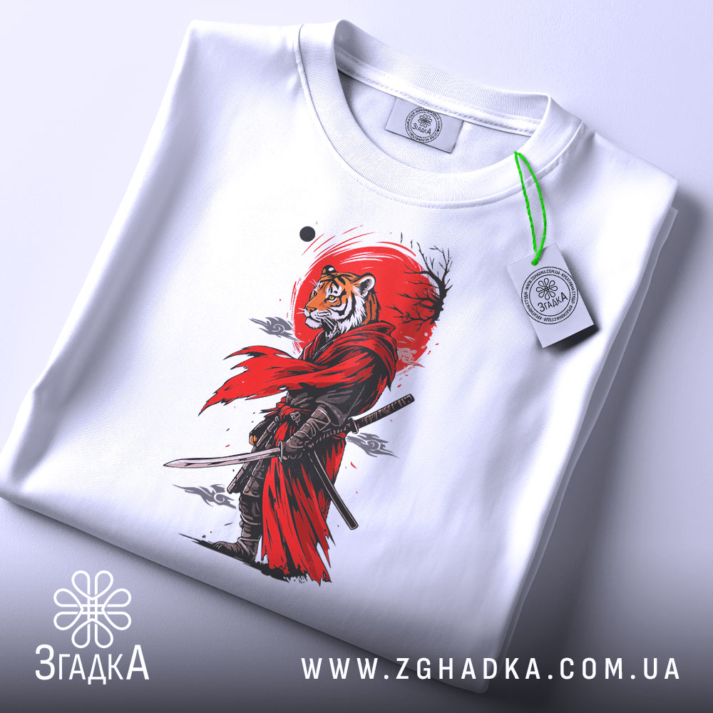 Футболка Тигр самурай воїн, біла, зображена на столі, фон світло-сірий, принт з тигром у червоному плащі, на футболці є етикетка з зеленим шнурком.