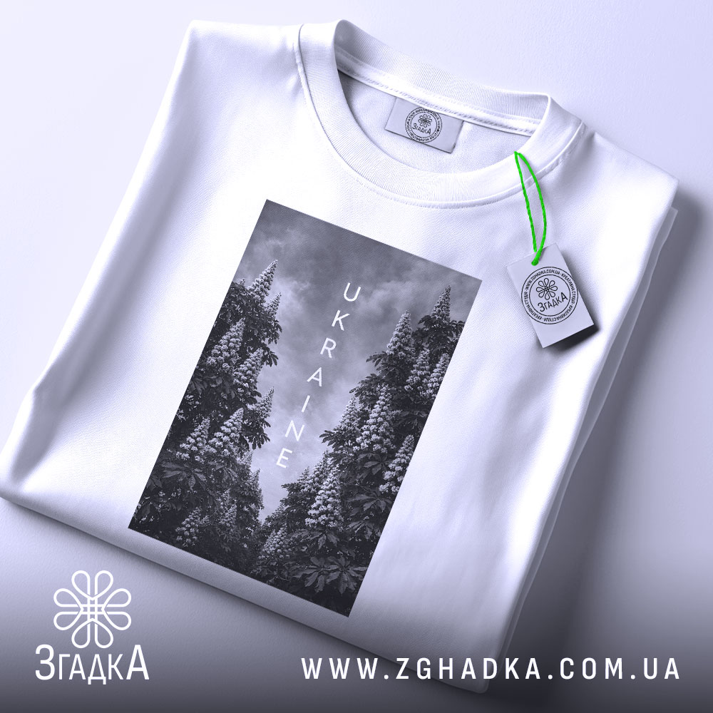 Футболка Ukraine білого кольору, складена на поверхні, з видимим етикеткою, фон світло-сірий, принт з чорнобілими зображеннями дерев та написом 'UKRAINE' по центру.