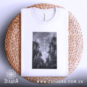 Футболка Ukraine білого кольору, складена на кошику з плетеного матеріалу, фон світло-сірий, принт з чорнобілими зображеннями дерев та написом 'UKRAINE' по центру.