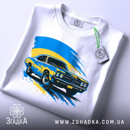 Футболка українське авто, біла, складена на столі, фон світло-сірий, принт з автомобілем жовтого кольору на синьому фоні з жовтими мазками, на футболці є етикетка.