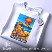 Футболка український настрій, біла, складена на столі, фон світло-сірий, принт з котом у сонцезахисних окулярах, що сидить на шезлонгу з напоєм, на фоні пляжу та вибуху, з етикеткою на комірці.