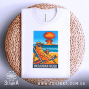 Футболка український настрій, біла, складена на плетеному кошику, фон світло-сірий, принт з котом у сонцезахисних окулярах, що сидить на шезлонгу з напоєм, на фоні пляжу та вибуху.